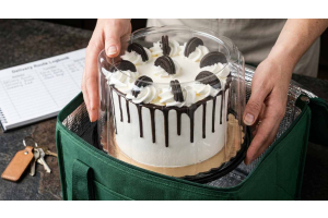 A professional baker placing a chocolate drip cake in a clear plastic cake dome container into an insulated green delivery bag, demonstrating the professional logistics of a bakery delivery fee.